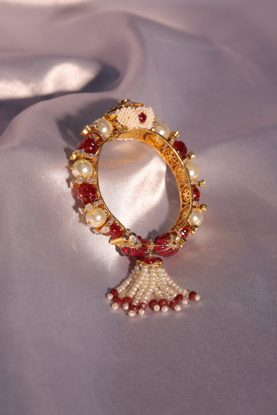 A gold-plated bangle adorned with pink polki, pearls, and ruby-colored stones, featuring a tassel design.