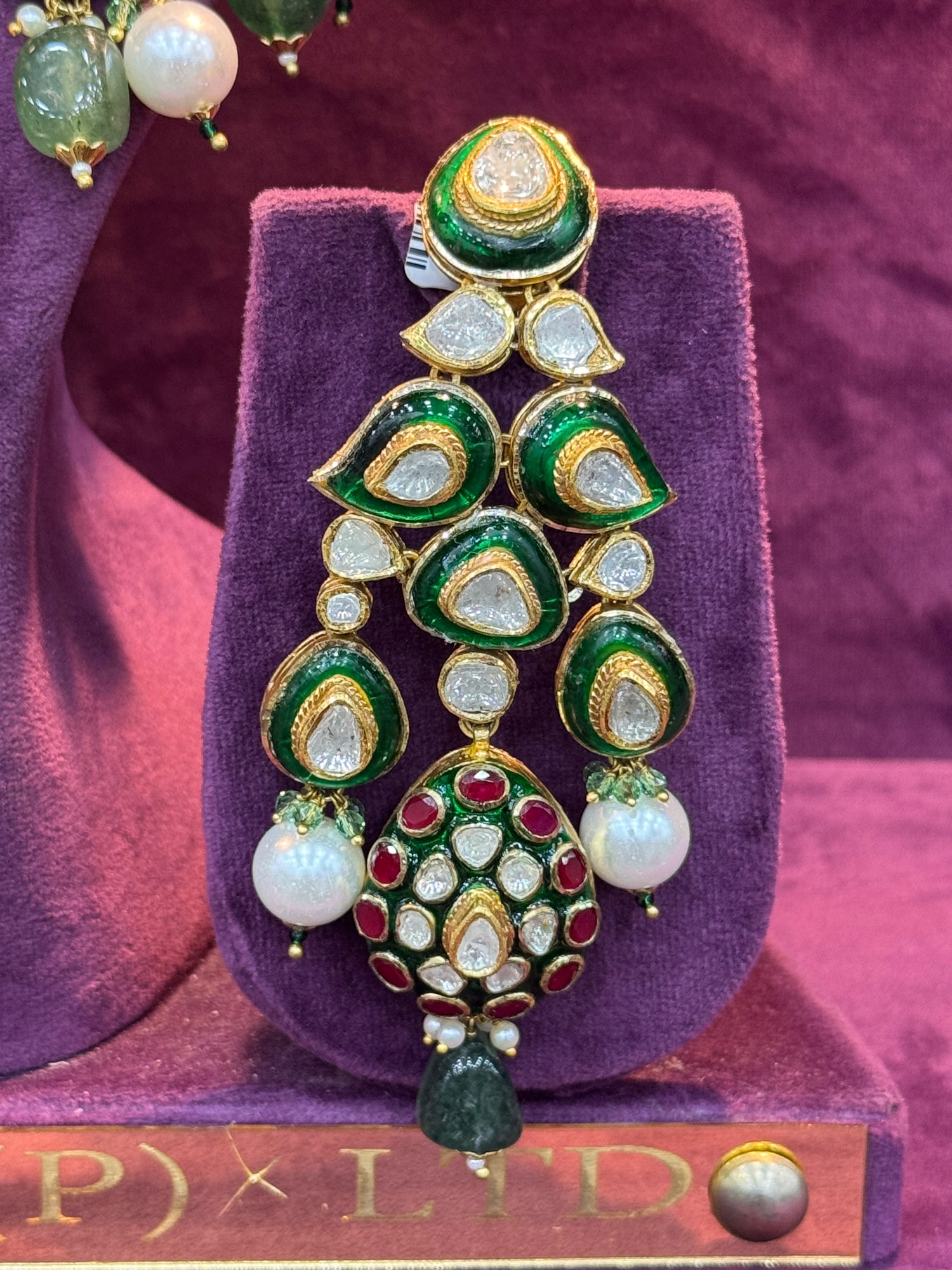 Ornate earrings with green, gold, and white stones on a purple stand with &