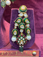 Ornate earrings with green, gold, and white stones on a purple stand with &
